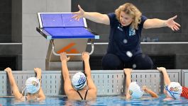 Otylia Jędrzejczak instruuje dzieci podczas zajęć nauki pływania. Siedzi na brzegu basenu przy słupku startowym, pokazując rękami ruchy, a młodzi pływacy w czepkach słuchają jej wskazówek, trzymając się krawędzi basenu.